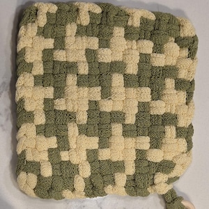 Pode incluir: Um pegador de panela quadrado, tecido, com um padrão xadrez de fio verde-oliva e creme. Uma pequena alça com um anel de madeira é presa a um canto. O pegador de panela é projetado para uso na cozinha.