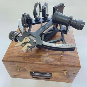 Op de afbeelding: Een zwarte sextant met de tekst "TAMAYA SEXTANT 1712" staat op een houten kist. Het instrument heeft lenzen, wijzerplaten en een vizier. De kist heeft een messing handvat en sluitingen.