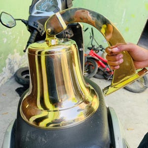 Puede incluir: Una campana de latón dorado brillante con un asa curva. La campana es cilíndrica con una base acampanada. El asa también es de latón y está unida a la parte superior de la campana.