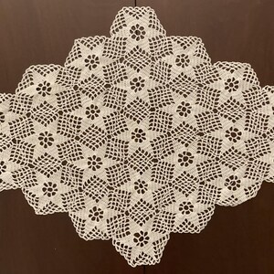 May include: Cream-colored, diamond-shaped crochet doily. The doily features a repeating floral pattern within a geometric design. The edges are scalloped. The doily is displayed on a dark brown wooden surface.