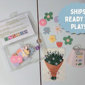 May include: A clear plastic case contains a game with flower-shaped pieces, dice, and cards. The game includes a card with a bouquet illustration and a small booklet. Various colorful flower cutouts are scattered around the case. A blue sign says "SHIPS READY TO PLAY!"