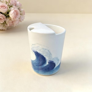 May include: White ceramic travel mug with a blue wave design. The mug has a white lid with a small opening. The wave design is painted in shades of blue and white, creating a dynamic and artistic look.
