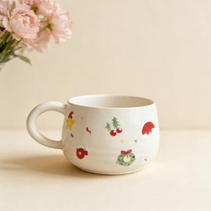 Puede incluir: Una taza de cerámica blanca con forma redondeada y un asa grande. La taza está decorada con ilustraciones coloridas con temática navideña, que incluyen una estrella, cerezas, un gorro de Papá Noel y una corona. La taza está sobre un fondo neutro.