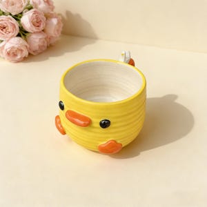 May include: A yellow ceramic planter shaped like a duck, with orange beak and feet, and black eyes. The planter has a ribbed texture and a white interior. A small bouquet of pink roses is in the background.