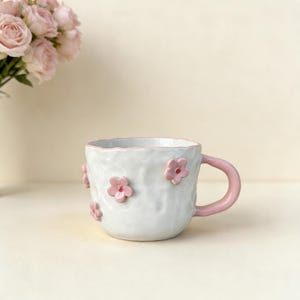 Op de afbeelding: Witte keramische mok met een getextureerd oppervlak en roze bloemaccenten. De mok heeft een roze handvat en is versierd met verschillende roze bloemen. Een boeket roze rozen staat op de achtergrond.