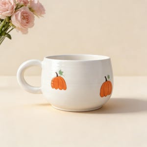 May include: White ceramic mug with a rounded shape and a large handle. The mug is decorated with two orange pumpkin illustrations with green leaves. A bouquet of pink roses is in the background.