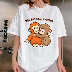 Puede incluir: Camiseta blanca con una ilustración de dibujos animados de dos monos abrazándose. Los monos están representados en tonos naranja y marrón. El texto "YOU ARE NEVER ALONE" está impreso en letras marrones encima de la ilustración.