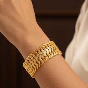 Puede incluir: Un brazalete dorado con un diseño tejido y texturizado. El brazalete es ancho y se envuelve alrededor de la muñeca. La joya está hecha de un material metálico brillante. El brazalete es una pieza llamativa.