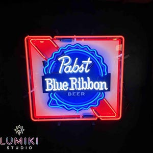 Op de afbeelding: Een neonreclame voor Pabst Blue Ribbon bier. Het bord toont een blauw en wit logo met de woorden "Pabst Blue Ribbon BEER" in een gestileerd lettertype. Het bord is ingekaderd in een rood-wit frame, met een zwarte achtergrond.