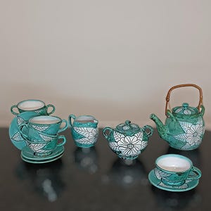 Pode incluir: Um conjunto de chá de cerâmica turquesa com desenhos florais brancos. O conjunto inclui chávenas, pires, um creme, um açucareiro e uma chaleira com uma pega de bambu. O conjunto de chá é exibido numa superfície escura.