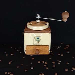 Pode incluir: Moinho de café Peugeot vintage, creme, com gaveta e manivela de madeira. O moinho, com o logótipo Peugeot, está sobre um fundo preto salpicado de grãos de café. A manivela é visível.