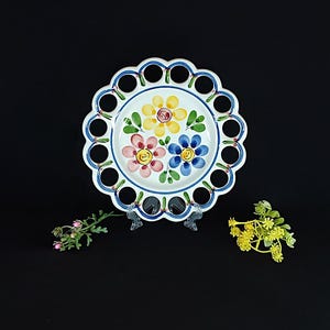 May include: A white ceramic plate with a scalloped edge and decorative cutouts. The plate features hand-painted flowers in yellow, pink, and blue. A blue border with cutouts surrounds the plate. The plate is displayed on a clear stand.