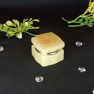 Puede incluir: Una pequeña caja de joyería cuadrada de color amarillo claro con un diseño floral en la tapa. La caja tiene una banda dorada alrededor del centro. El fondo es negro, con gemas decorativas redondas y transparentes dispersas y ramitas de vegetación.