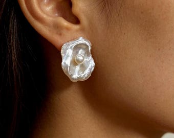 May include: A stud earring featuring an irregular, organic-shaped white shell-like component with a lustrous, iridescent finish. A single, small, round white pearl is centered within the concave, textured surface of the shell-like piece.