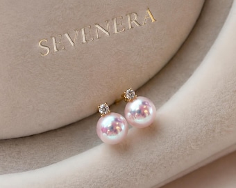 May include: A pair of stud earrings featuring round, iridescent pink pearls topped with small, clear, faceted stones set in gold-toned metal. The earrings are positioned near a beige display case with the gold-embossed text SEVENERA.
