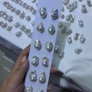 May include: A vertical white display card featuring six pairs of irregular, lustrous white baroque pearls arranged in two columns. The pearls exhibit a high-gloss finish with subtle iridescent undertones, and many loose pearls are scattered nearby.