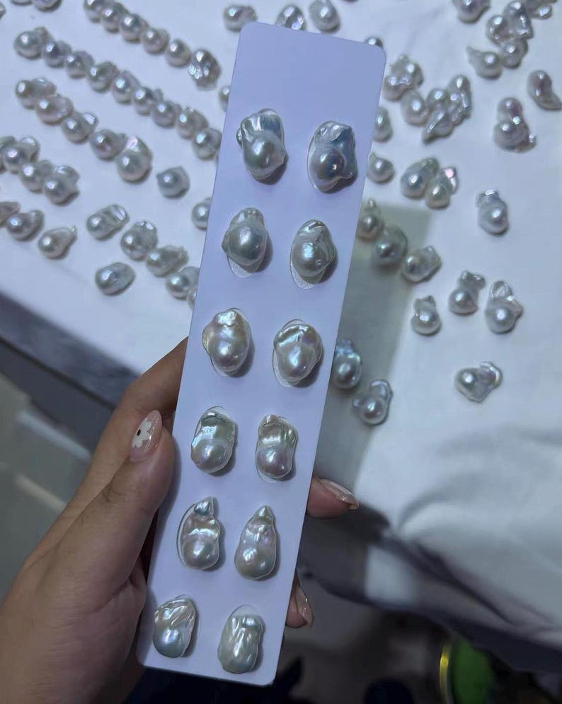 May include: A vertical white display card featuring six pairs of irregular, lustrous white baroque pearls arranged in two columns. The pearls exhibit a high-gloss finish with subtle iridescent undertones, and many loose pearls are scattered nearby.