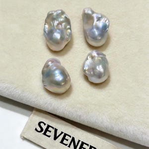 May include: Four irregular, baroque-shaped pearls with a lustrous, iridescent finish displaying soft pink, blue, and silver tones. The pearls are arranged in a square formation above a fabric tag featuring the black text SEVENERA.