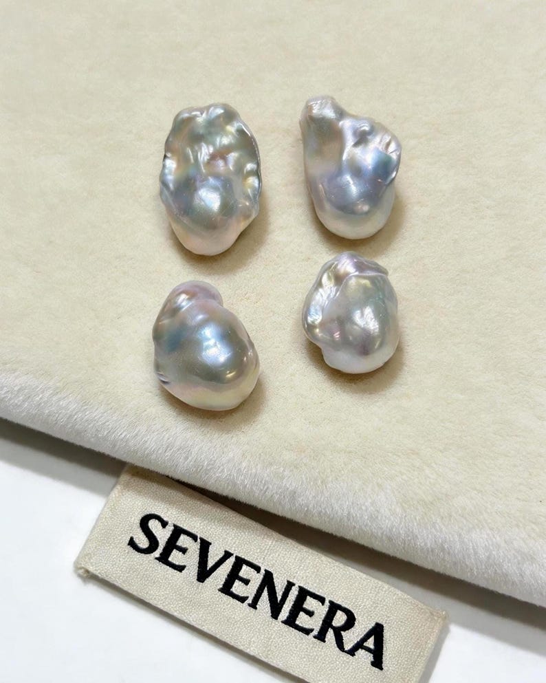 May include: Four irregular, baroque-shaped pearls with a lustrous, iridescent finish displaying soft pink, blue, and silver tones. The pearls are arranged in a square formation above a fabric tag featuring the black text SEVENERA.