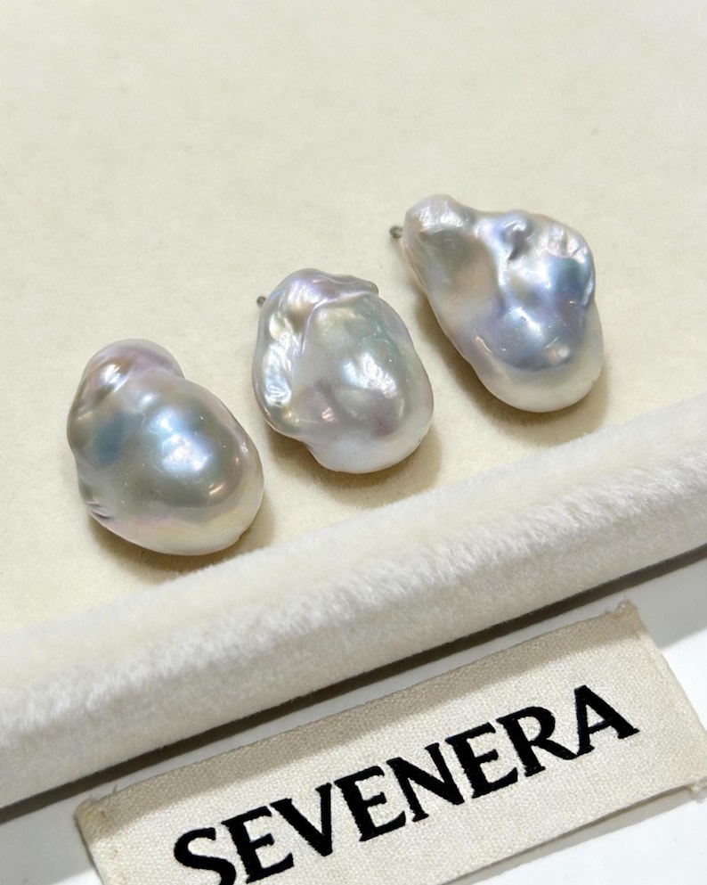 May include: Three irregular, lustrous baroque pearls with iridescent white and silver tones. Each pearl features a metallic post at the top for attachment. A small fabric tag with the black text SEVENERA is visible in the lower right corner.