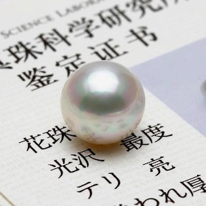 May include: A spherical white pearl with a subtle iridescent sheen resting on a document featuring black Japanese and English text. The pearl has a smooth, reflective surface and sits centrally on the paper, which includes the text Pearl Science Laboratory.