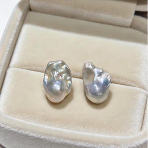 May include: Two irregular, baroque-shaped pearls with a lustrous, iridescent surface displaying subtle hues of silver, cream, and soft pink. The pearls have a textured, organic appearance with a high-gloss finish, resting side by side.