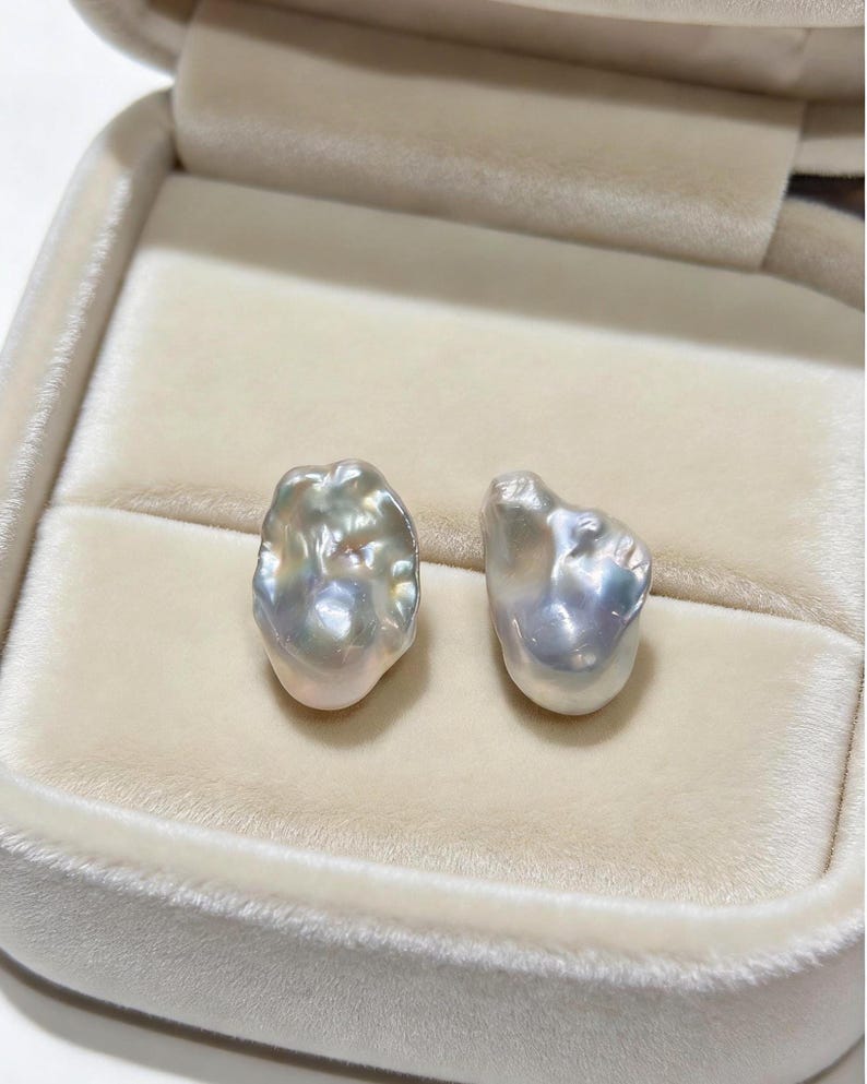 May include: Two irregular, baroque-shaped pearls with a lustrous, iridescent surface displaying subtle hues of silver, cream, and soft pink. The pearls have a textured, organic appearance with a high-gloss finish, resting side by side.