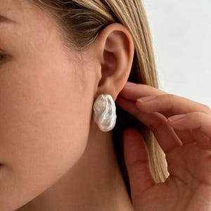 May include: A single white baroque pearl earring with an irregular, organic shape and a high-luster, iridescent surface. The pearl features a textured, bumpy finish and is secured directly against the earlobe, showcasing its natural, non-spherical form.