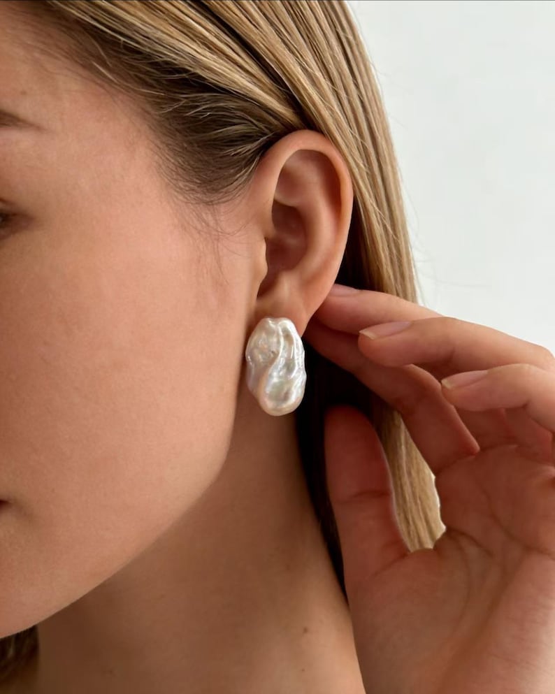 May include: A single white baroque pearl earring with an irregular, organic shape and a high-luster, iridescent surface. The pearl features a textured, bumpy finish and is secured directly against the earlobe, showcasing its natural, non-spherical form.
