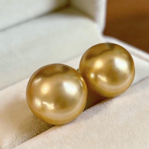 Puede incluir: Dos grandes y brillantes perlas doradas descansan dentro de un joyero de terciopelo blanco. Las perlas tienen una forma esférica suave y un brillo cálido y radiante. Un accesorio lujoso.