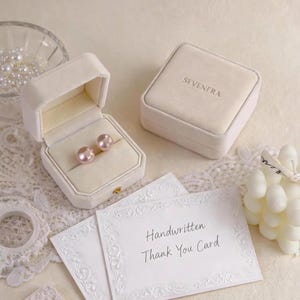 May include: Two beige velvet jewelry boxes, one open containing two round pink pearl earrings and one closed with the text SEVENERA, alongside two white cards with the text Handwritten Thank You Card and a small glass bowl of loose pearls.