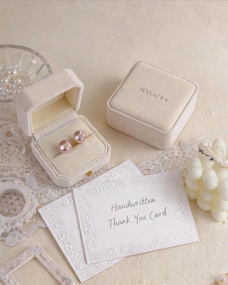 May include: Two beige velvet jewelry boxes, one open containing two round pink pearl earrings and one closed with the text SEVENERA, alongside two white cards with the text Handwritten Thank You Card and a small glass bowl of loose pearls.