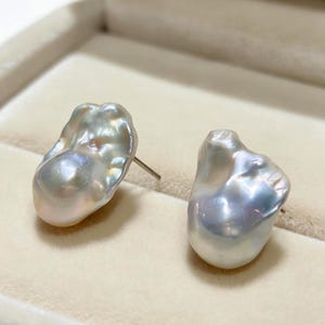 May include: A pair of stud earrings featuring irregular, baroque-shaped pearls with a lustrous, iridescent white and silver finish. Each pearl displays a unique, organic surface texture with subtle pink and blue undertones, attached to a metal post.
