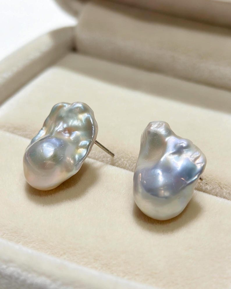 May include: A pair of stud earrings featuring irregular, baroque-shaped pearls with a lustrous, iridescent white and silver finish. Each pearl displays a unique, organic surface texture with subtle pink and blue undertones, attached to a metal post.
