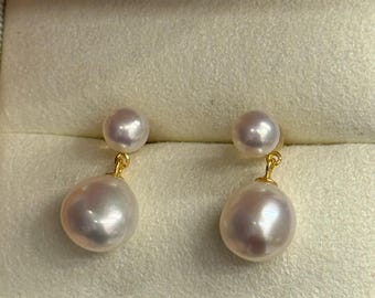 May include: A pair of pearl earrings. Each earring features a small, round pearl stud connected to a larger, irregularly shaped pearl drop. The earrings are suspended from gold-colored hooks, presented in a beige jewelry box.