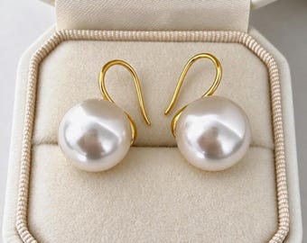 May include: A pair of gold hook earrings featuring large, round, lustrous white pearls. The earrings are displayed inside a cream-colored, fabric-lined jewelry box with a structured, square-shaped design and a soft, velvet-like interior lining.