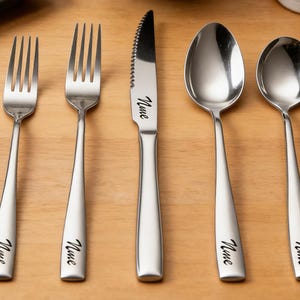 Puede incluir: Un juego de cinco cubiertos de acero inoxidable: dos tenedores, un cuchillo y dos cucharas. Cada pieza tiene un diseño sencillo y moderno con la palabra "Nine" grabada en el mango. Los cubiertos están colocados sobre una superficie de madera marrón claro.