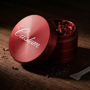 May include: A red metal grinder with the word "Custom" in white script on the lid. The grinder is open, revealing the grinding teeth. A small, clear plastic scraper is next to the grinder on a wooden surface.