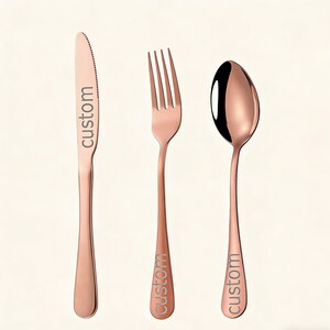 Könnte beinhalten: Roségoldenes Besteck-Set mit Messer, Gabel und Löffel. Jedes Teil trägt das Wort "custom" auf dem Griff eingraviert. Das Messer hat eine gezahnte Klinge. Der Löffel hat eine abgerundete Schale.