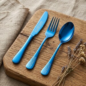 May include: A set of three blue stainless steel utensils: a knife, fork, and spoon. Each piece has the word "custom" printed on the handle. The utensils are arranged on a wooden cutting board with dried lavender sprigs.