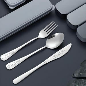Könnte beinhalten: Ein Edelstahl-Besteckset mit Gabel, Löffel und Messer. Jedes Teil ist mit dem Wort "CCUSTOM" graviert. Das Besteck liegt auf einer dunklen Oberfläche, mit einem grauen Etui im Hintergrund.