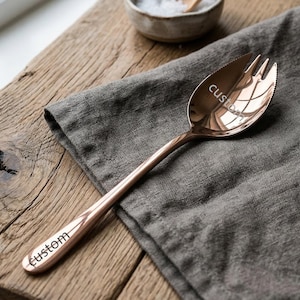 Może przedstawiać: Miedziana łyżka do serwowania o unikalnym designie, z końcówką w kształcie widelca i napisem "custom" wygrawerowanym na trzonku i główce łyżki. Łyżka spoczywa na szarej lnianej serwetce, umieszczonej na drewnianej powierzchni.