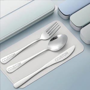 May include: A stainless steel flatware set includes a fork, spoon, and knife, each with the word "CCUSTOM" engraved on the handle. The set is displayed on a light gray placemat, with a light blue and white case in the background.