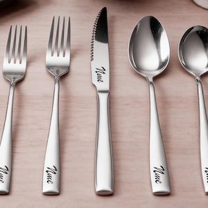 May include: A set of five stainless steel silverware pieces: two forks, a knife, and two spoons. Each piece has a simple, elegant design with the word "Nine" etched on the handle. The silverware is arranged on a light brown surface.