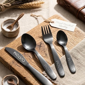 Könnte beinhalten: Ein Set aus vier mattschwarzen Besteckteilen: ein Messer, ein Löffel, eine Gabel und ein Teelöffel, jeweils mit dem Wort "CUSTOM" graviert. Das Besteck liegt auf einem Holzschneidebrett mit einer kleinen Schale Salz und einem Holzlöffel. Ein Etikett trägt die Aufschrift "Hand-Finished Quality".