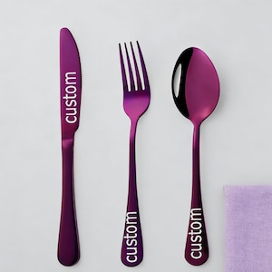 May include: A set of three pieces of purple flatware: a knife, a fork, and a spoon. The knife and fork have the word "custom" printed vertically on the handles. The spoon has a reflective surface. A purple napkin is in the background.