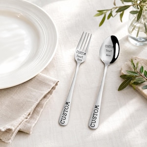 May include: A silver-toned metal fork and spoon set featuring engraved text. The fork head reads CUSTOM Text Here with CUSTOM on the handle. The spoon bowl reads CUSTOM Text Here with CUSTOM on the handle. Both utensils have a polished finish.