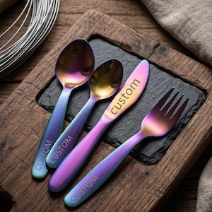 May include: A set of iridescent flatware, including a spoon, a smaller spoon, a knife, and a fork. The word "CUSTOM" is engraved on each piece. The flatware is displayed on a black slate and a wooden serving board.