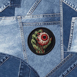 May include: A denim patch featuring a green hand holding a red eyeball with blood dripping down. The patch is circular with a black border and is set against a background of various denim pieces.