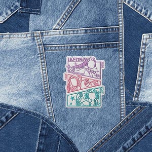 May include: Close-up of a denim patchwork design. A colorful patch with three illustrated figures and the text "HUNTRA" is sewn onto the denim. The patch features the names "RUMI", "MIRA", and "ZOEY".